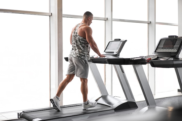 Modelo de fitness culturista atlético muscular corriendo gimnasio caminadora cerca de una ventana grande