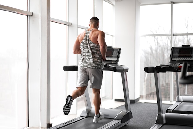 Modelo de fitness culturista atlético muscular corriendo gimnasio caminadora cerca de una ventana grande