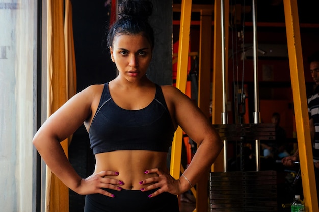 Modelo de fitness latinoamericano en sujetador y calzas con aspecto cansado de sudor en la frente en el gimnasio oscuro