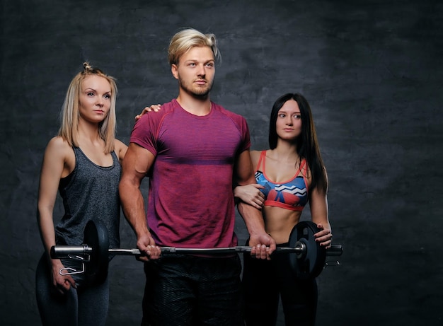 Modelo de fitness trío de mujeres rubias y morenas delgadas y un hombre sostiene una barra posando sobre un fondo gris.