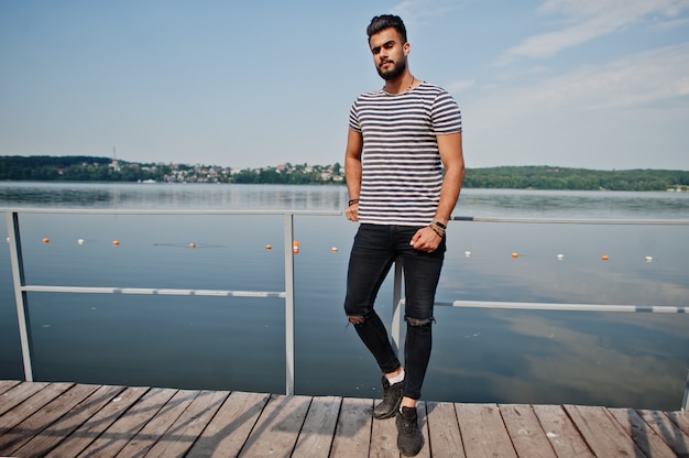 Modelo de hombre de barba árabe alto guapo en camisa despojado planteado al aire libre en el muelle del lago. Chico árabe de moda.