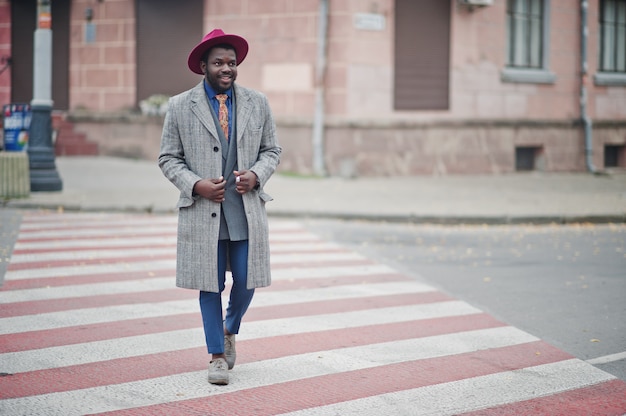 Modelo de hombre elegante con abrigo gris, corbata y sombrero rojo caminando en el cruce peatonal