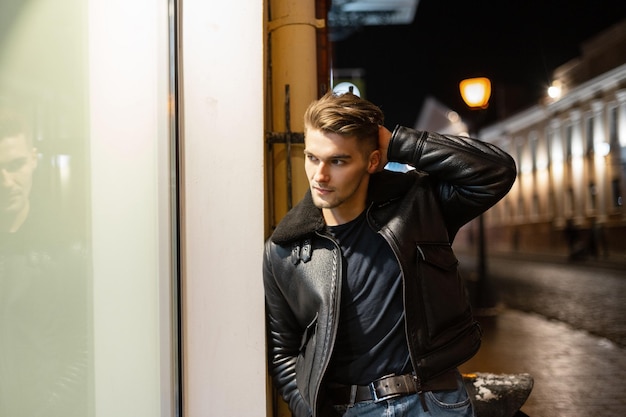 Modelo de hombre joven y apuesto de moda con peinado en una elegante chaqueta de cuero negro caminando en una ciudad nocturna y de pie cerca de un escaparate