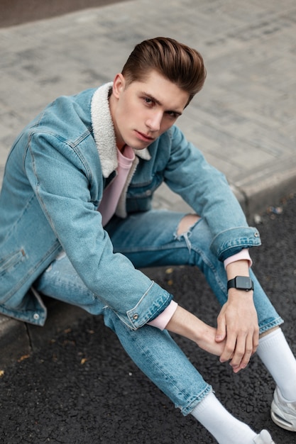 Modelo de hombre joven europeo con peinado en chaqueta de mezclilla azul de moda en jeans rasgados con estilo en zapatillas blancas descansando sobre baldosas cerca de la carretera. Chico atractivo en ropa casual juvenil en la calle en la ciudad.