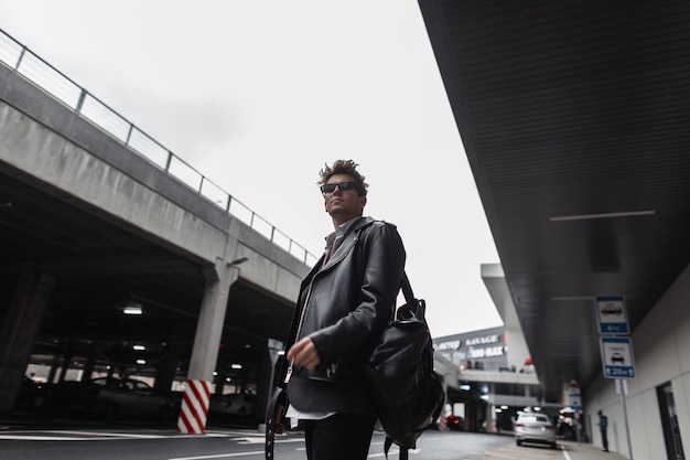 Modelo de hombre joven de moda estadounidense en una chaqueta de cuero negro de moda de gran tamaño juvenil con peinado con mochila se encuentra cerca de un edificio moderno en la ciudad. Chico guapo hipster en gafas de sol al aire libre.