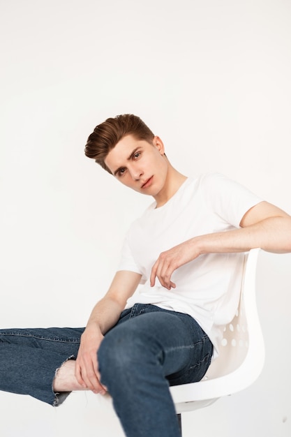 Modelo de hombre joven muy agradable con peinado en camiseta blanca de moda en jeans de moda azul posando en silla junto a la pared vintage en el interior. Chico guapo en ropa casual elegante descansa en el estudio.