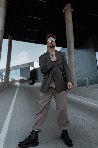 Modelo de hombre de negocios de moda guapo en elegante traje gris con botas y gafas de sol sobre fondo urbano