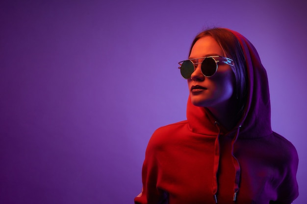 Modelo joven posando en una luz de neón de moda. Mujer en gafas de sol redondas y sudadera con capucha con efectos de luz de colores. Estilo disco. Filtro de gel.