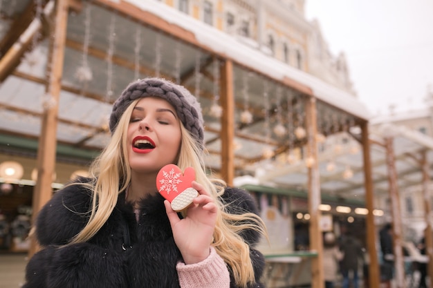 Modelo loira atraente com olhos fechados segurando um biscoito de gengibre saboroso na decoração leve na praça em Kiev