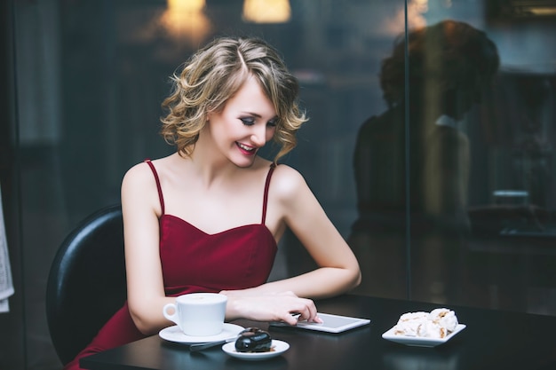 Modelo loira linda mulher em um macacão vermelho na moda e elegante com uma xícara de café e tablet no restaurante