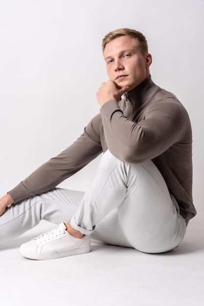 Modelo loiro caucasiano com um suéter marrom sobre um fundo branco, sentado no chão com um olhar sério