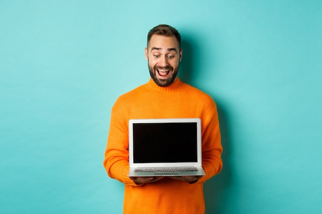 Modelo masculino adulto empolgado mostrando a tela do laptop, demonstrando promoção on-line e feliz