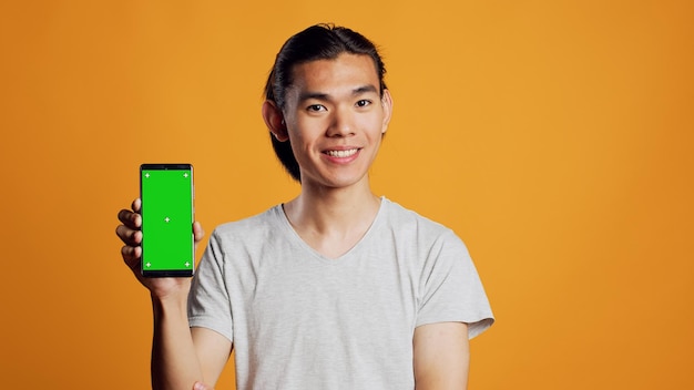 Modelo masculino asiático que muestra un teléfono inteligente con pantalla verde en la cámara, mirando el espacio de copia de la clave de croma en el estudio. Joven persona moderna que usa teléfono móvil con pantalla de maqueta aislada.