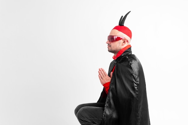 Modelo masculino caucásico en traje rojo negro de halloween y gafas de sol con sombrero y cuernos rezando sobre fondo blanco.