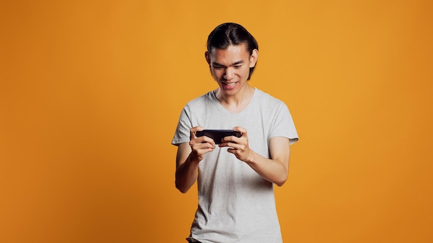 Modelo masculino feliz jogando videogame no smartphone, usando a competição de jogos online para se divertir no estúdio. Jovem asiático desfrutando de um jogo para celular no telefone, jogo de desafio de atirador.