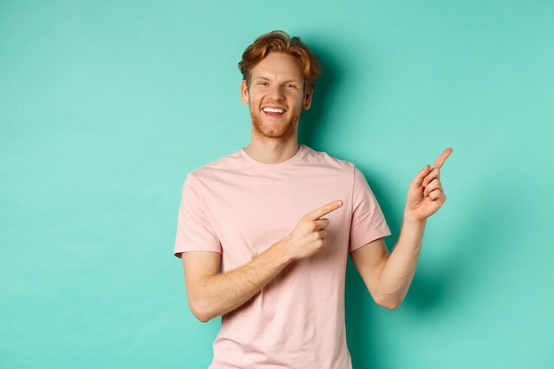 Modelo masculino guapo con cabello rojo desordenado que muestra publicidad en el espacio de la copia, apuntando a la esquina superior derecha y sonriendo feliz, de pie sobre fondo turquesa