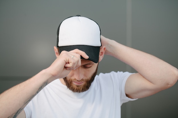 Modelo masculino guapo hipster con barba con camiseta blanca en blanco y una gorra de béisbol con espacio para su logotipo o diseño en estilo urbano casual