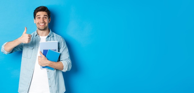 Foto modelo masculino jovem feliz segurando cadernos e mostrando o polegar para cima satisfeito sorrindo e recomendando o curso