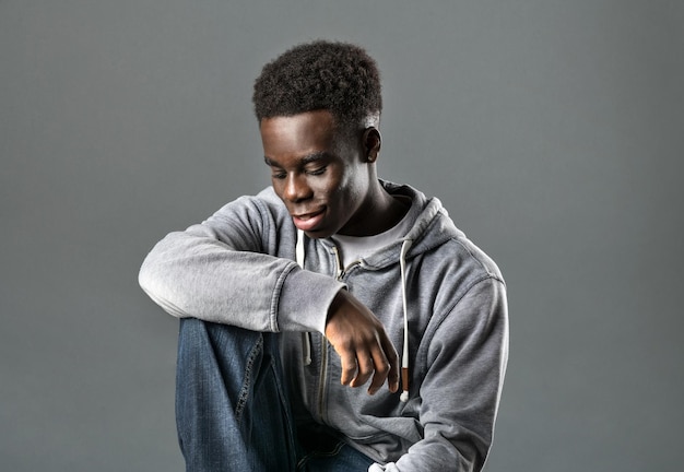 Modelo masculino negro joven positivo con cabello afro oscuro en una sudadera con capucha de moda y jeans sonriendo y mirando hacia abajo mientras está sentado contra el fondo gris en el estudio