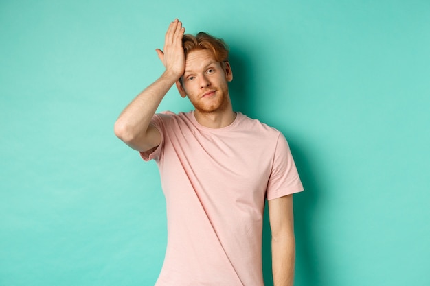 Modelo masculino ruiva irritada e incomodada, mostrando o gesto facepalm, em pé sobre o fundo da casa da moeda.