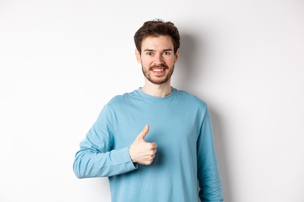 Modelo masculino sonriente en camisa casual mostrando el pulgar hacia arriba, aprobar y recomendar el producto, elogiar la excelente elección, de pie sobre fondo blanco.