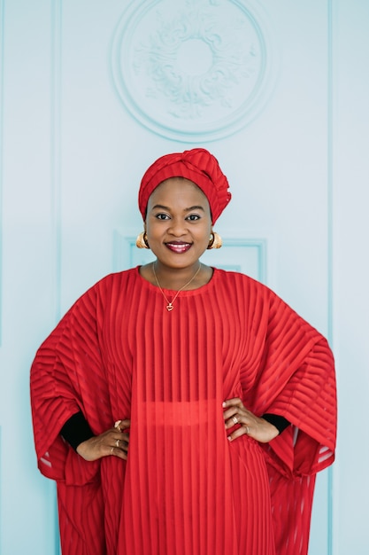 Modelo de moda africana en estilo africano tradicional con elegante traje rojo, pañuelo en la cabeza, aretes y maquillaje sobre fondo de puerta azul claro en estudio.