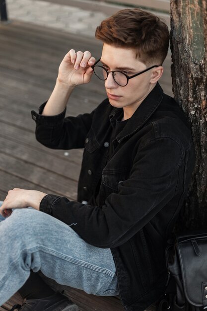 Modelo de moda apuesto joven en ropa de mezclilla de moda endereza gafas vintage al aire libre