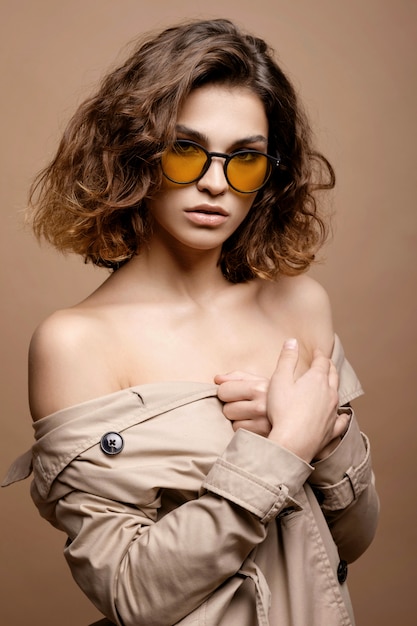 Modelo de moda de belleza con piel limpia y cabello rizado con capa de biege elástica, modelo con gafas de moda, hombros descubiertos