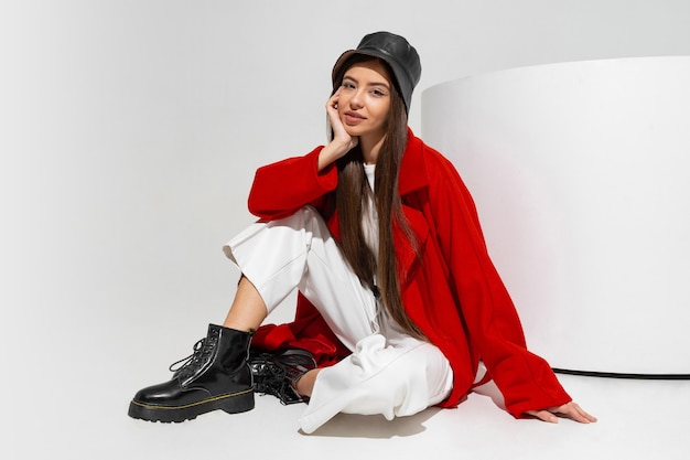 Modelo de moda en elegante sombrero, abrigo rojo y botas posando en la pared blanca