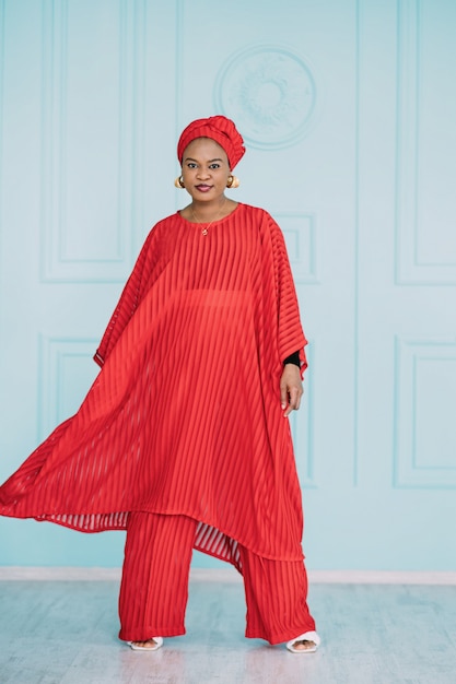 Modelo de moda femenina africana que parece feliz posando en el estudio. Disparo de longitud completa de hermosa mujer en elegante ropa roja sobre fondo azul claro.