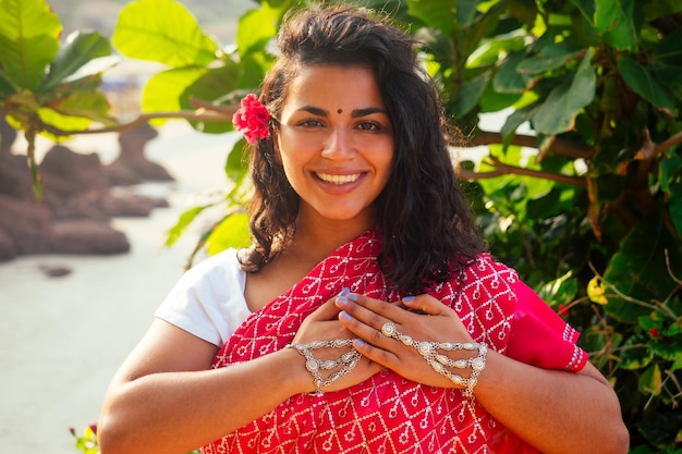 Modelo de moda femenina india sonrisa de dientes blancos con flor en el pelo rizado en traje tradicional de la india sari de boda rojo posando cerca de árboles tropicales en un paraíso playa mar oceanwellness balneario