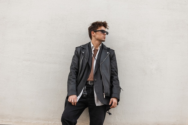 Modelo de moda joven con una elegante chaqueta de cuero negro en una camisa de jeans vintage con una mochila con gafas de sol descansa cerca de una pared gris en la ciudad. Hipster de chico americano urbano al aire libre.