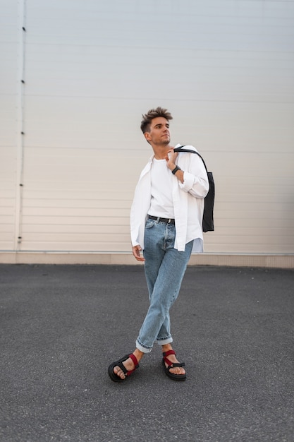 Modelo de moda joven estadounidense en jeans de moda con una elegante camisa en sandalias de moda de cuero rojo con bolso de tela negro posa cerca de un edificio blanco. Chico moderno. Nueva colección de verano de moda masculina.