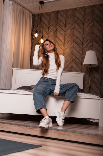 Modelo de moda de una mujer joven elegante en un top elegante blanco en jeans de moda posando en la cama en el dormitorio. Chica pelirroja en ropa de moda está descansando en la habitación con un interior moderno.