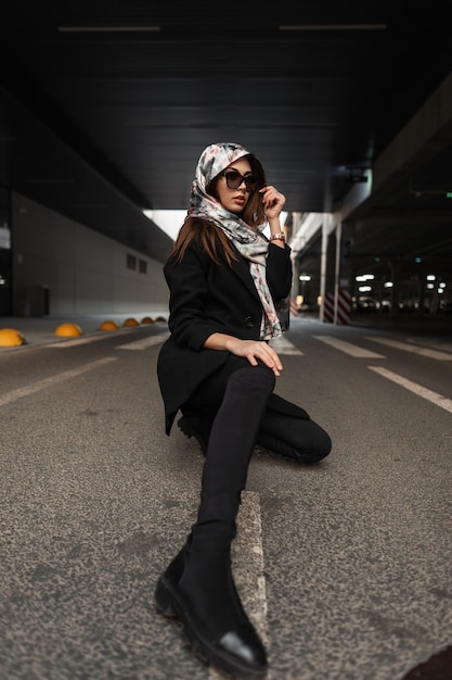 Modelo de moda mujer joven con elegantes gafas de sol en seda elegante pañuelo en la cabeza en un moderno abrigo negro en jeans juveniles con botas posando sentado en el asfalto en la ciudad. Chica de negocios se relaja en el asfalto.