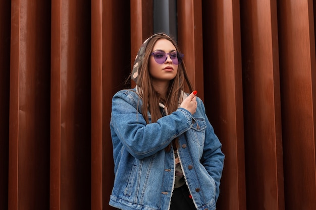 Modelo de moda mujer joven inconformista en gafas moradas de moda en chaqueta de mezclilla azul de gran tamaño con el pelo largo descansa cerca de la pared de metal en la calle. Modelo sexy chica fresca con estilo en la calle.