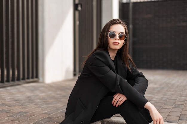Modelo de moda mujer joven con labios sensuales en elegantes gafas de sol en ropa negra de moda de la nueva colección de moda juvenil descansa cerca del edificio en la ciudad. Cool hipster chica hermosa sentarse al aire libre.
