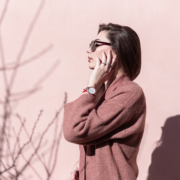 Modelo de moda de mujer joven muy de moda con hermoso cabello largo en elegantes gafas de sol en elegante abrigo de primavera se encuentra cerca de la pared rosa vintage y disfruta de la luz del sol brillante. Hermosa chica posando al aire libre