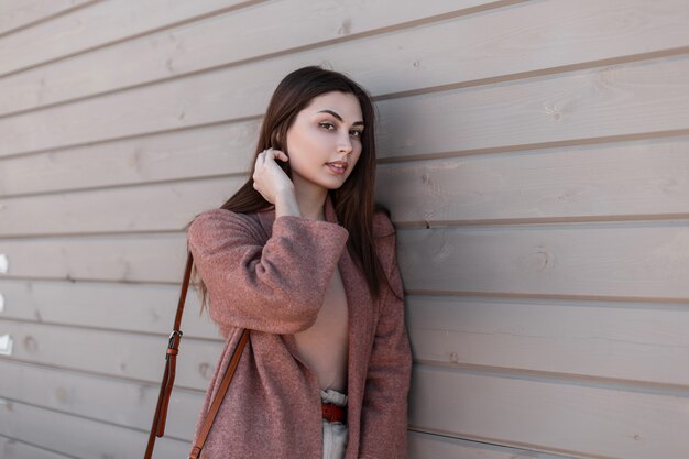 Modelo de moda de mujer joven urbana linda en ropa elegante de primavera posando cerca de edificio vintage de tablones. Bella y encantadora modelo de niña con estilo urbano endereza el cabello y disfruta de descansar cerca de la pared de madera al aire libre