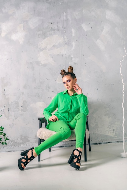 Modelo de moda profesional de tiro en estudio moderno. Una mujer elegante con traje verde se sienta en una silla de fondo gris. Marco vertical.