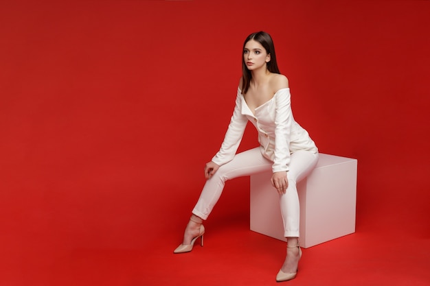 Modelo de moda se sienta en el cubo en pantalones de traje blanco Hermosa mujer joven Disparo de estudio Fondo rojo