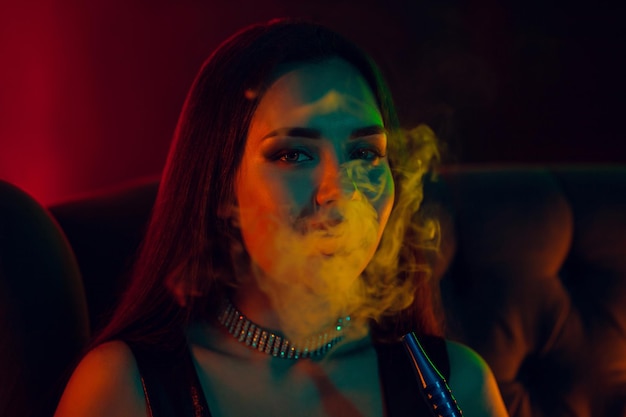 Modelo morena sexy está fumando una pipa de agua exhalando un humo en un club nocturno de lujo.