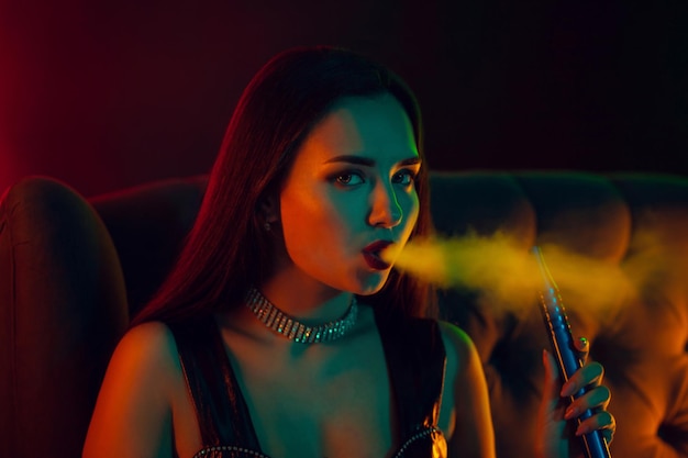 Modelo morena sexy está fumando um cachimbo de água, exalando uma fumaça em uma boate de luxo.