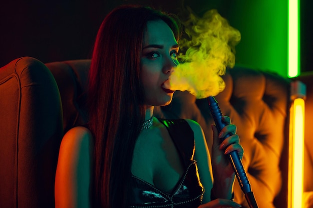 Modelo morena sexy está fumando um cachimbo de água, exalando uma fumaça em uma boate de luxo.
