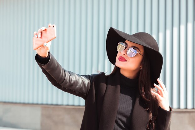 Modelo morena sonriente tomando selfie en teléfono móvil. Mujer lleva sombrero y gafas de sol