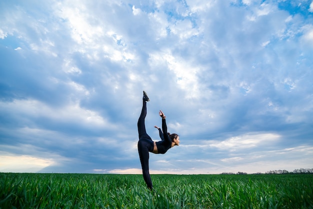 Modelo de la muchacha hermosa en la hierba verde hacer yoga. Una hermosa morena sobre un césped verde realiza elementos acrobáticos. gimnasta flexible en negro está haciendo ejercicios