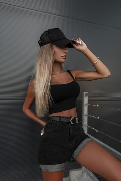 Modelo muito jovem, branca, com um boné preto na moda com shorts jeans pretos e uma blusa com uma pulseira de joias posando na rua perto de uma parede de metal escuro