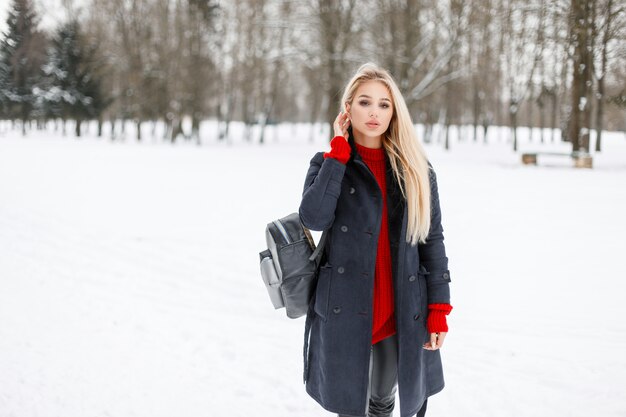 Modelo muito jovem em um casaco da moda com um suéter vermelho e uma bolsa estilosa caminhando no parque