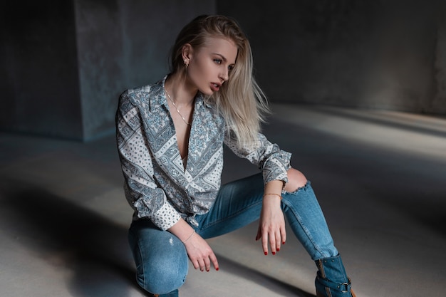 Modelo muito sexy de mulher jovem e bonita em uma camisa vintage com um padrão em jeans rasgados em sapatos elegantes, posando em um estúdio cinza em um dia ensolarado