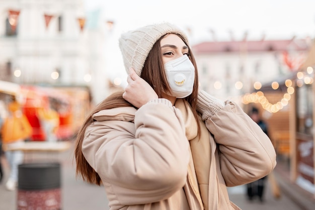 Modelo de mujer bastante joven y elegante con máscara médica protectora en ropa de invierno de moda con un sombrero de punto y chaqueta de plumas camina por la ciudad